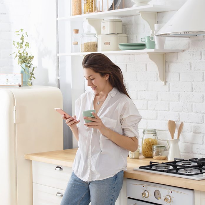 Young woman using smartphone leaning at kitchen table with coffee mug and organizer