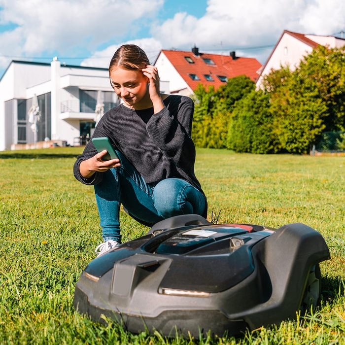 Young Woman Controlling Robot Lawn Mower In Summer Garden