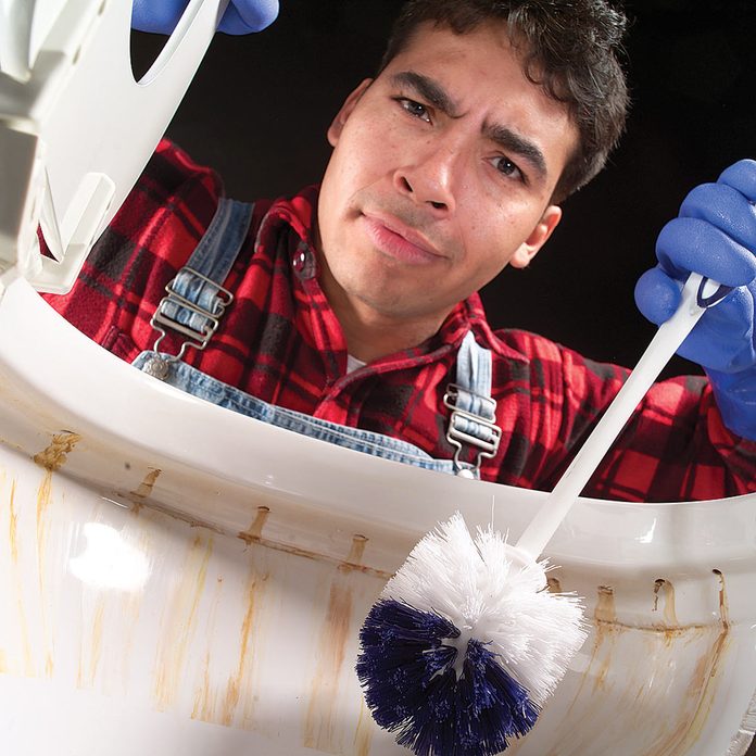 Man holds toilet brush above toilet with rust stains. 