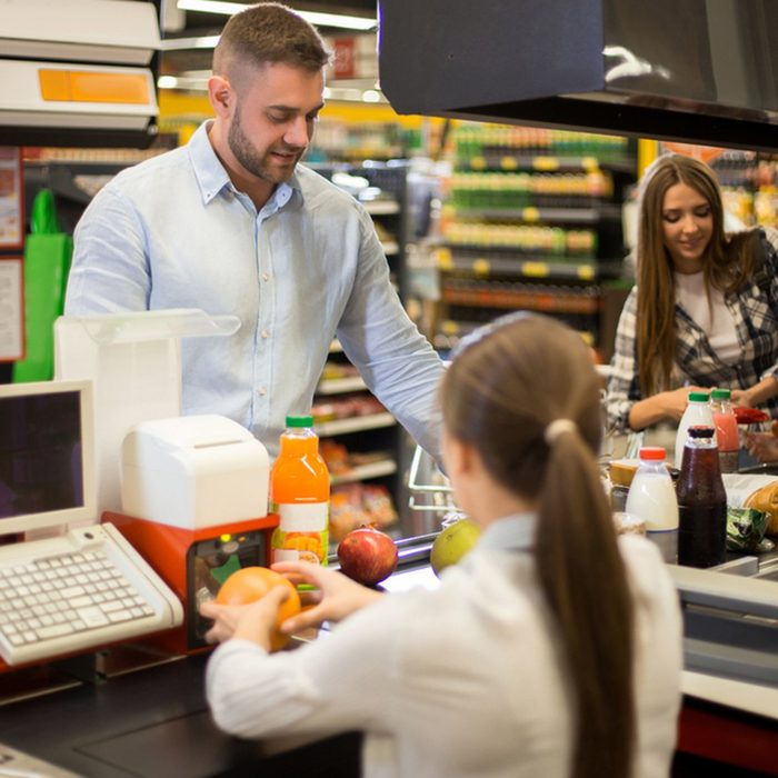 Portrait of smiling young man buying food in supermarket watching cashier scanning prices at cash desk and paying for groceries