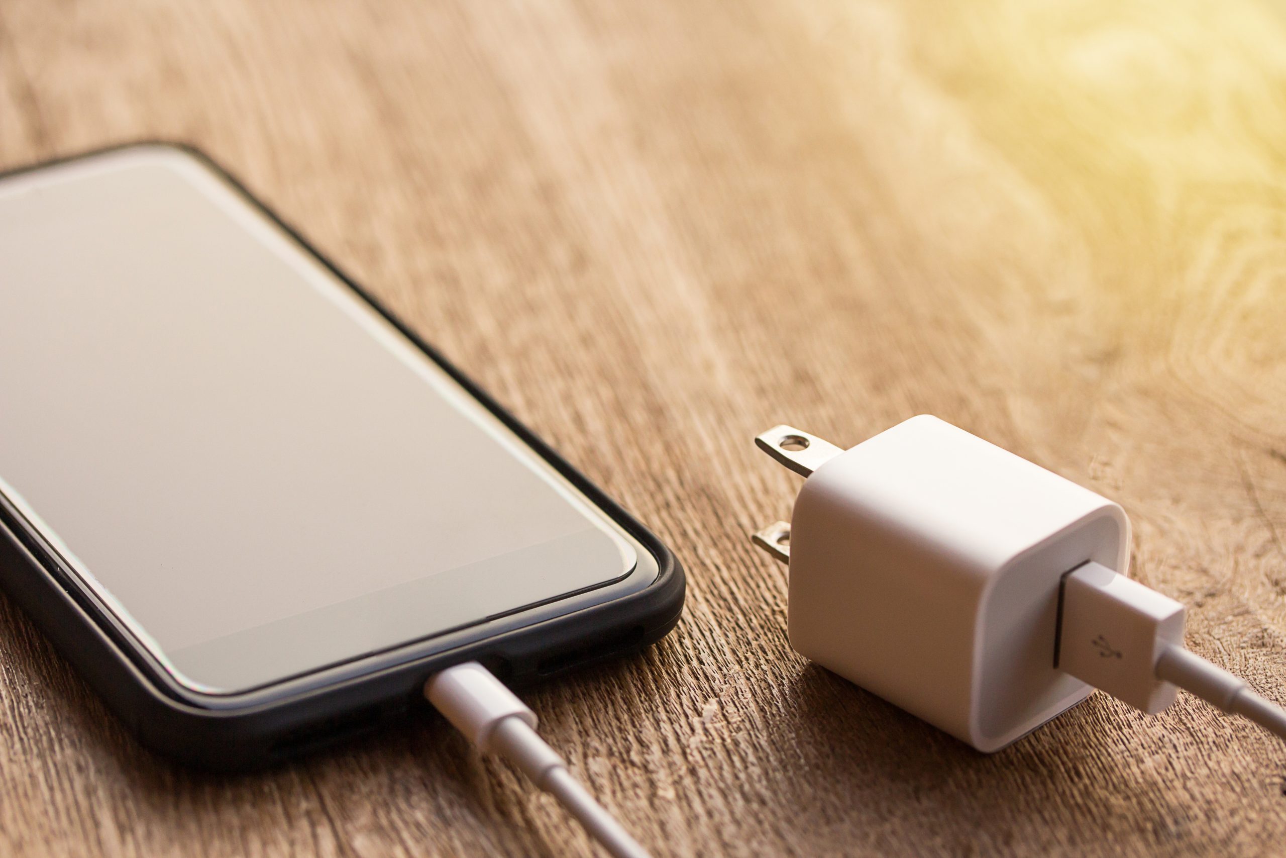 Close up of black smartphone charging battery with cable on wooden table and sunlight with copy space and blurry background. Vintage tone and soft light. Selective focus.