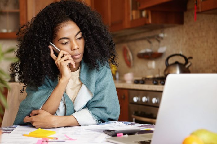 Sad African female with Afro hairstyle sitting in kitchen in front of laptop, talking on mobile phone to her husband, telling him that their family will be evicted soon because of non-payment for rent