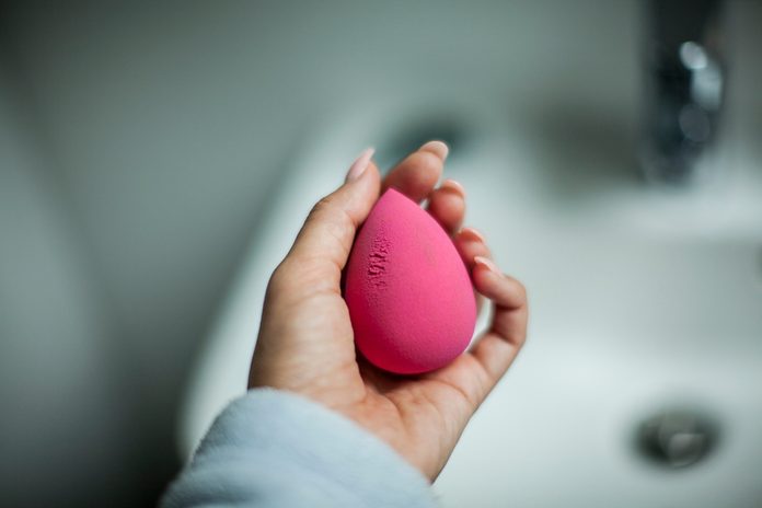 girl holds in her hand a pink rosa sponge for make-up, on the background there is a bright bathroom and a wash basin