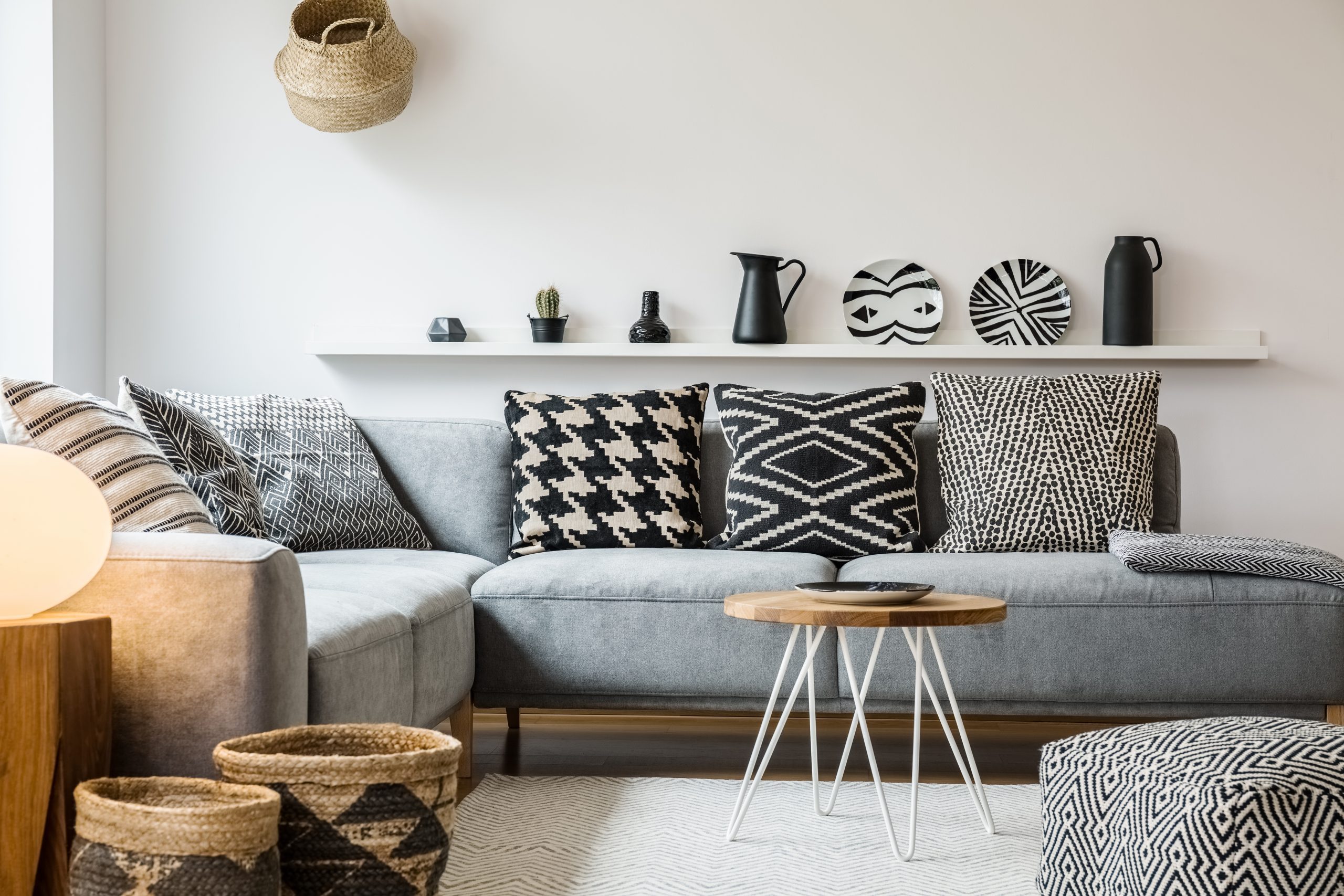 Patterned pillows on grey couch in modern living room interior with pouf and wooden table. Real photo