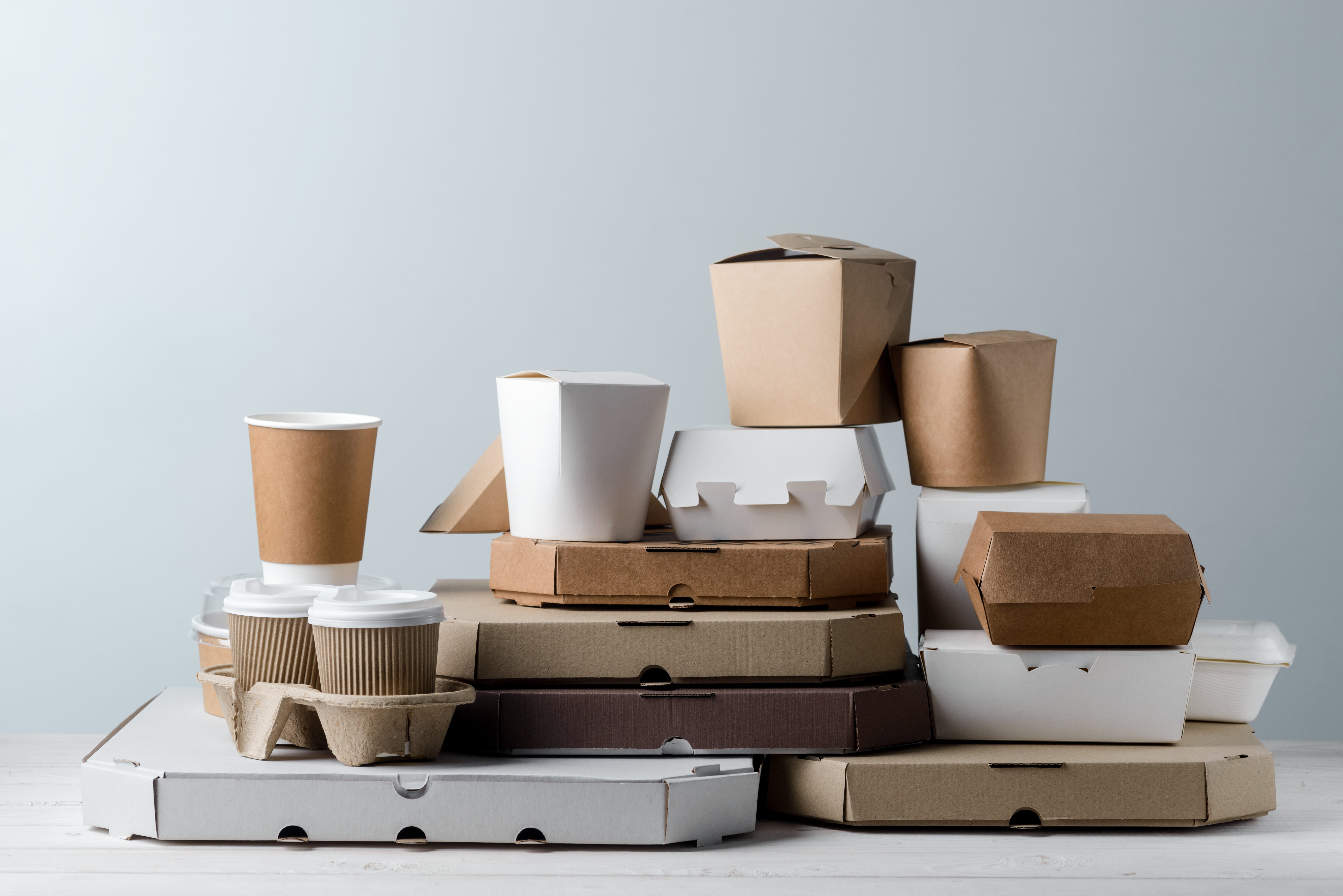 Assortment of pizza boxes, paper food containers, take-out coffee cups in holder, close-up. Light grey background, wooden surface. Food delivery.