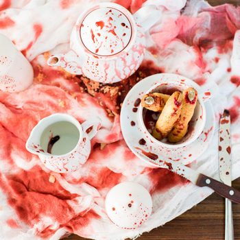 Bloody Halloween composition: high angle view of ceramic crockery in blood placed on dirty gauze, witch finger cookies in bloody cup