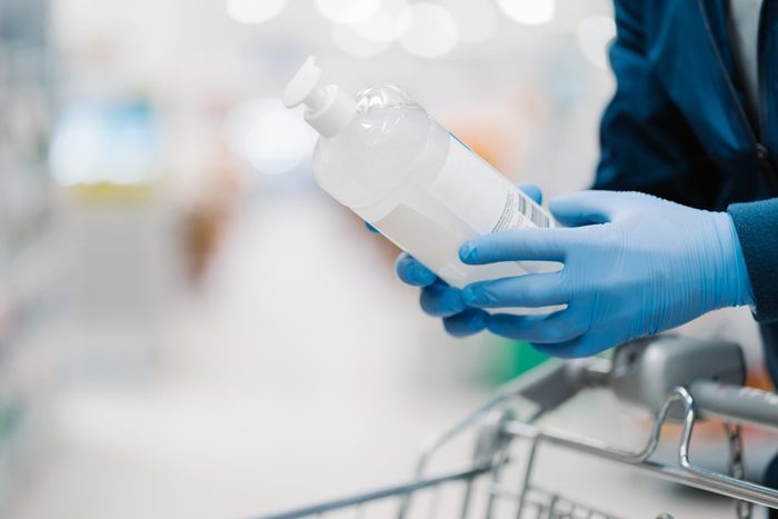 Unrecognizable person in medical gloves holds bottle of sanitizer gel, poses in shop, leans at shopping cart, buys product for coronavirus protection, blurred background. Disinfection, prevention