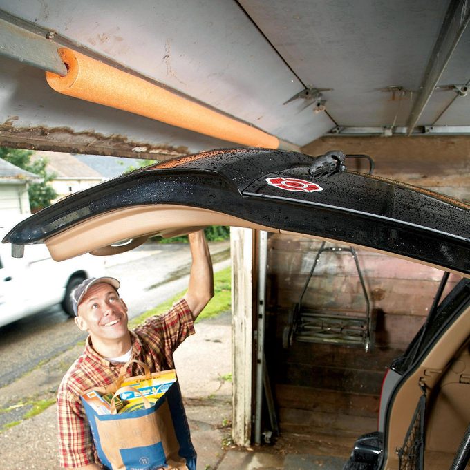 a man with groceries in his hands opening a car hatch door in a garage. The garage door with an orange pool noodle to protect the car's trunk door