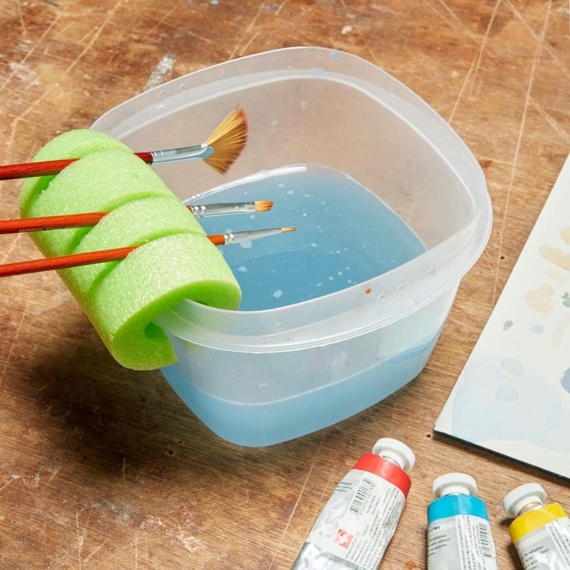 small paint brushes resting in a slits in a green pool noodle that has been placed on the edge of a plastic container that holds paint rinse water