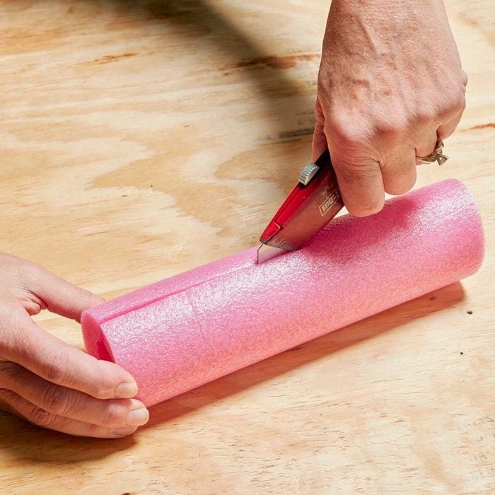 using a box cutter to cut a slit down the center of a small pink pool noodle