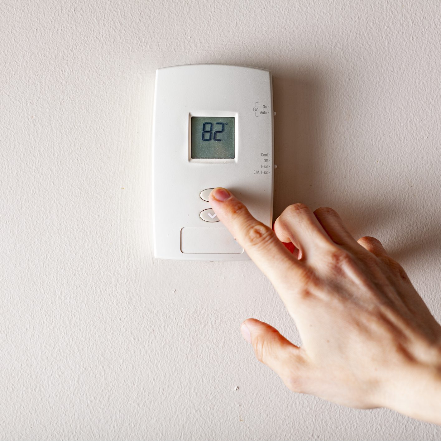 A woman is pressing the up button of a wall attached house thermostat with digital display showing the temperature