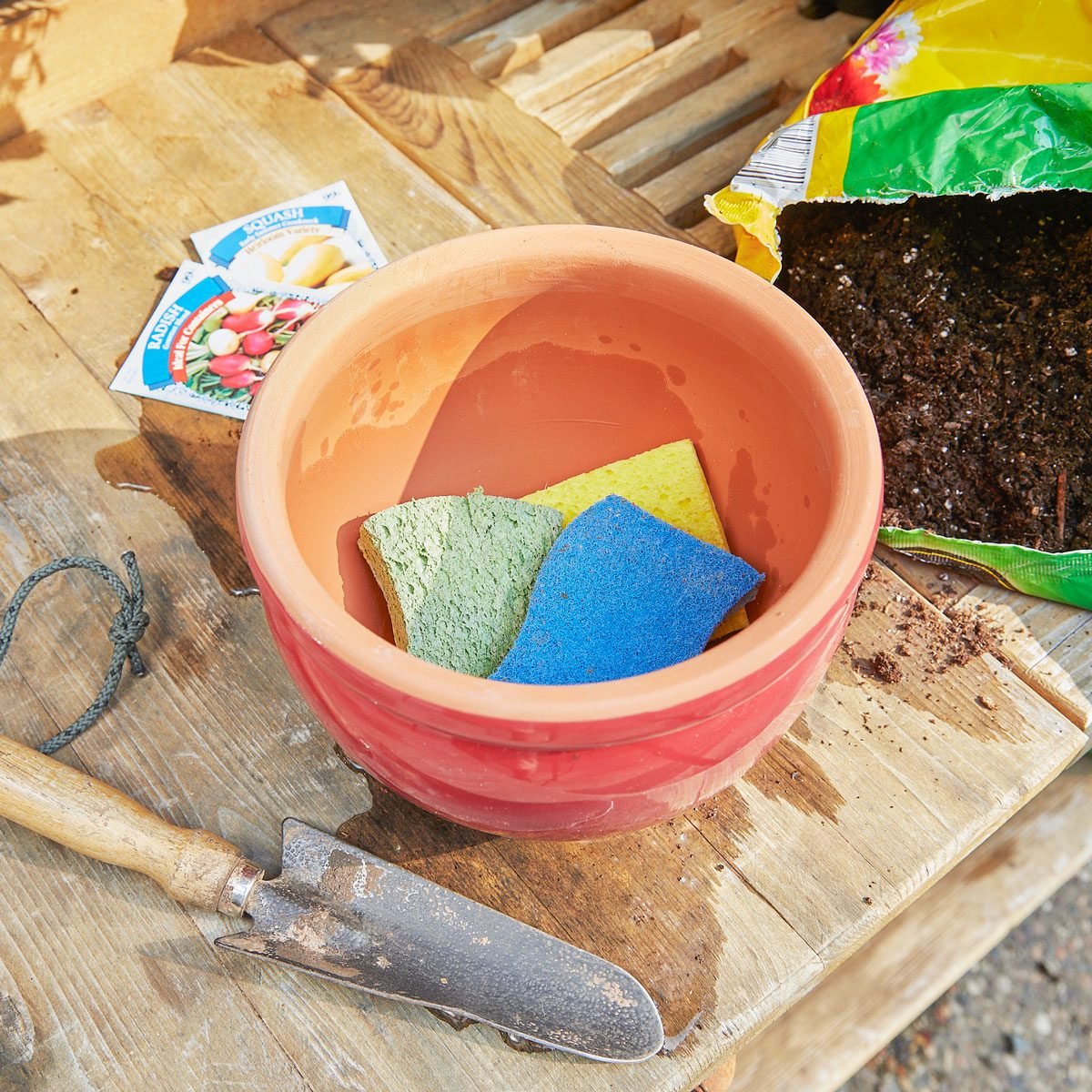 three spongs at the base of a terra cotta pot near potting soil, seed packets, and gardening shovel