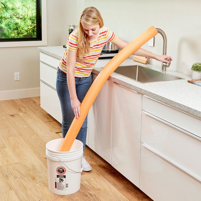 woman using a pool noodle to fill a bucket on the floor from the kitchen sink