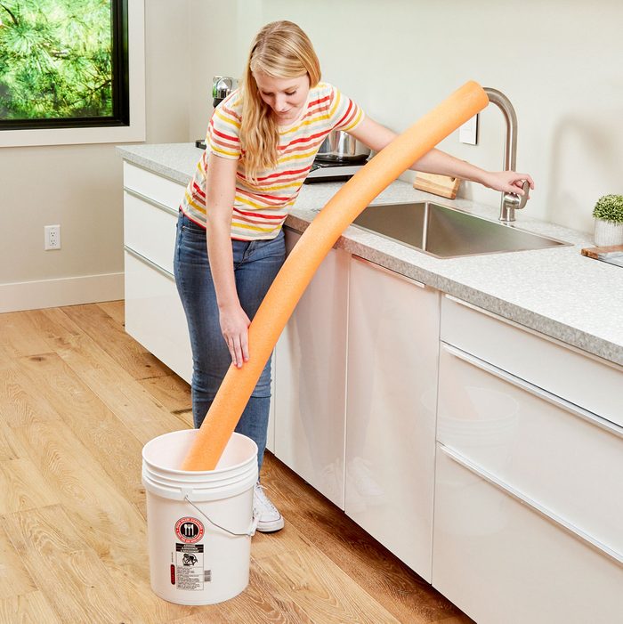 woman using a pool noodle to fill a bucket on the floor from the kitchen sink