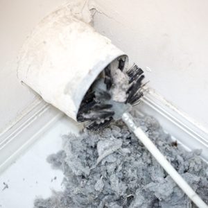 Dryer vent in a home being cleaned out with a round brush. There is a large pile of lint that has been removed from the vent on a white tiled floor. The walls and baseboards are white. The lint is gray