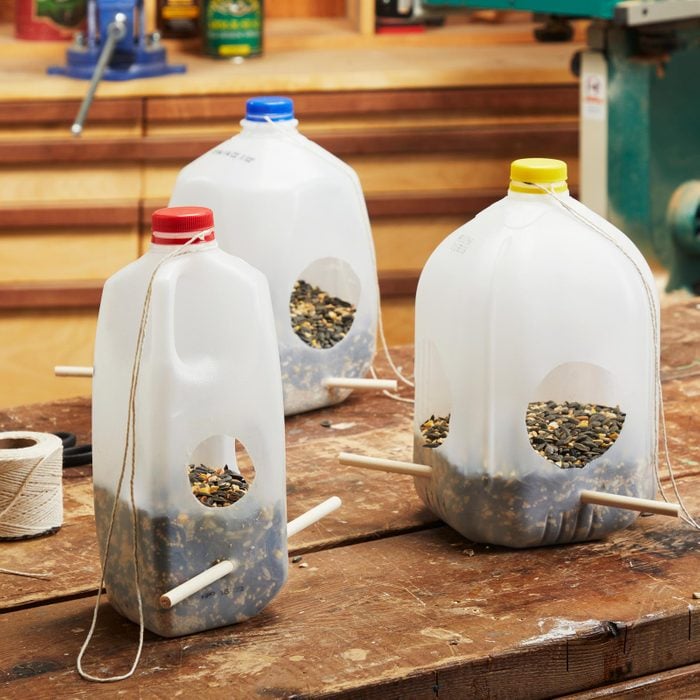 three Milk Jug Bird Feeders of different sizes filled with bird seed sitting on a work bench