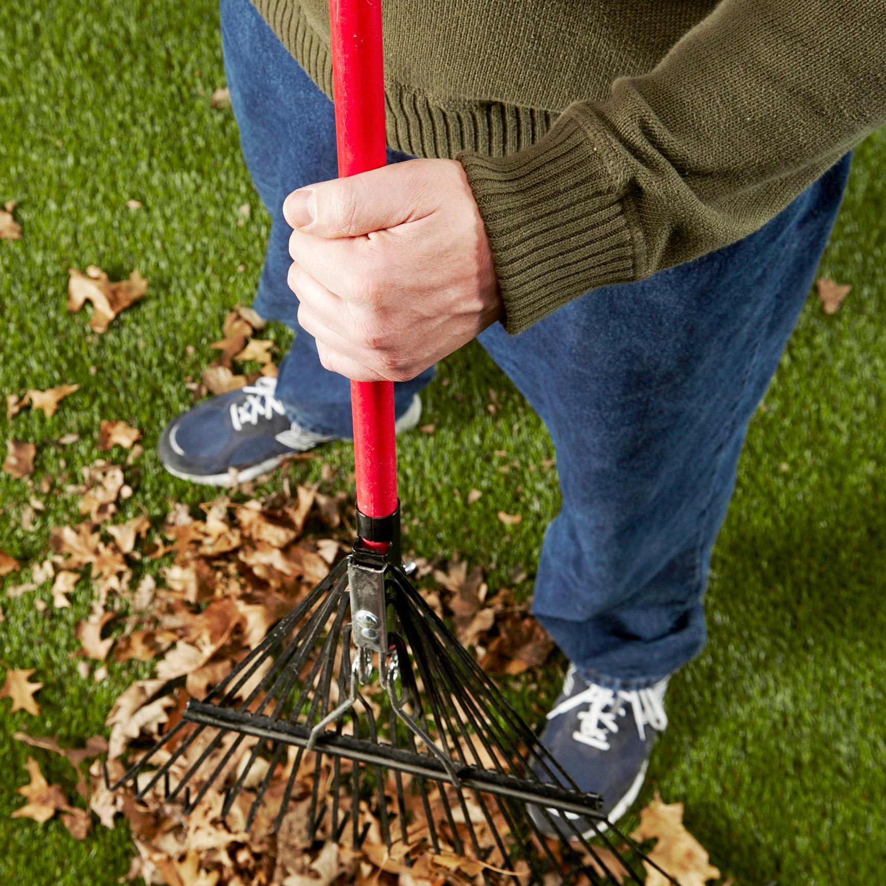 man in green sweater and blue jeans holding a red rake with fall leaves in the grass