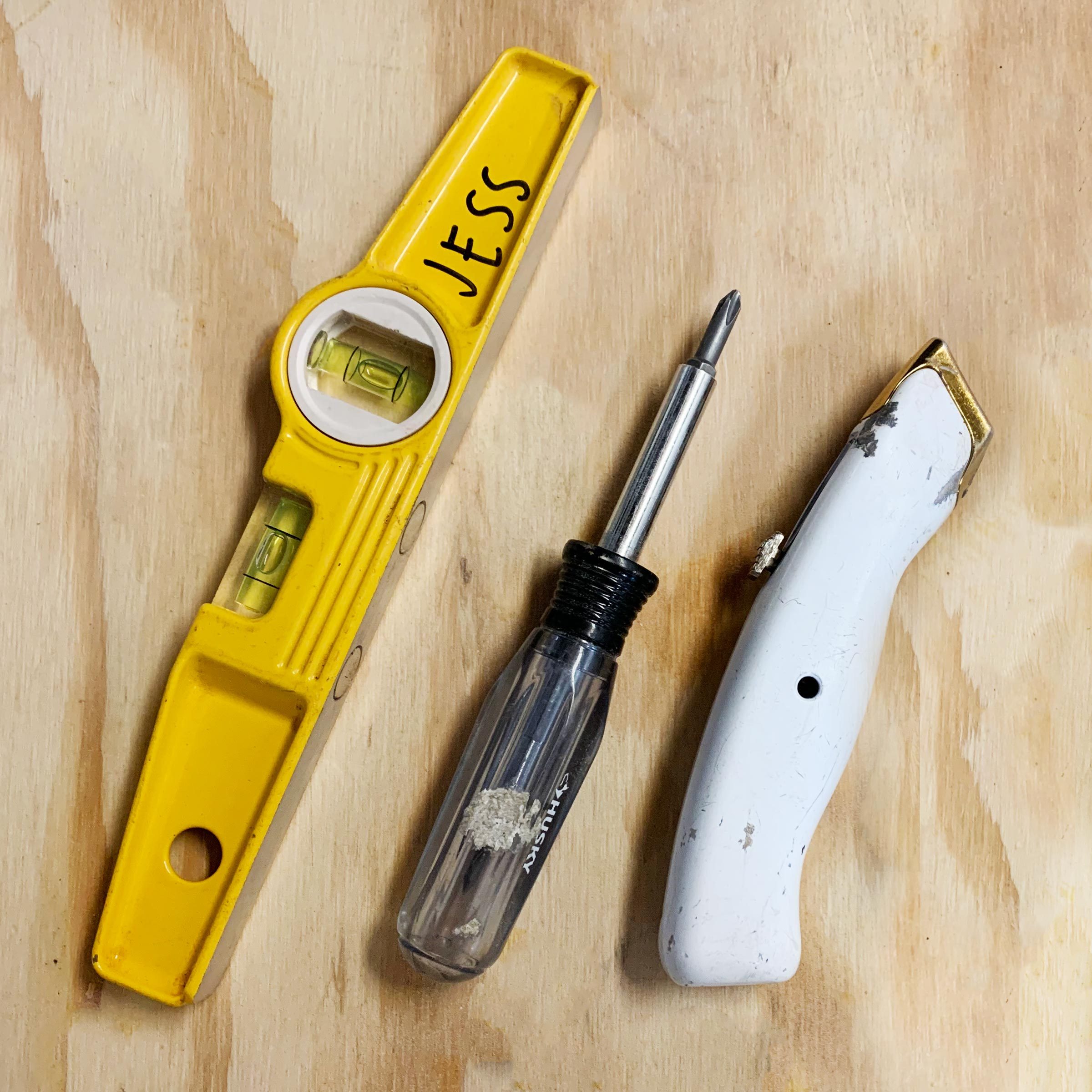 three tools lined up on a piece of plywood including a level, a screwdriver and a box cutter