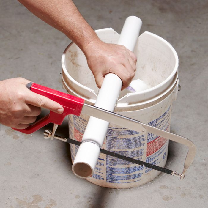 using a bucket to rest PVC pipe while using a hacksaw to cut the pipe