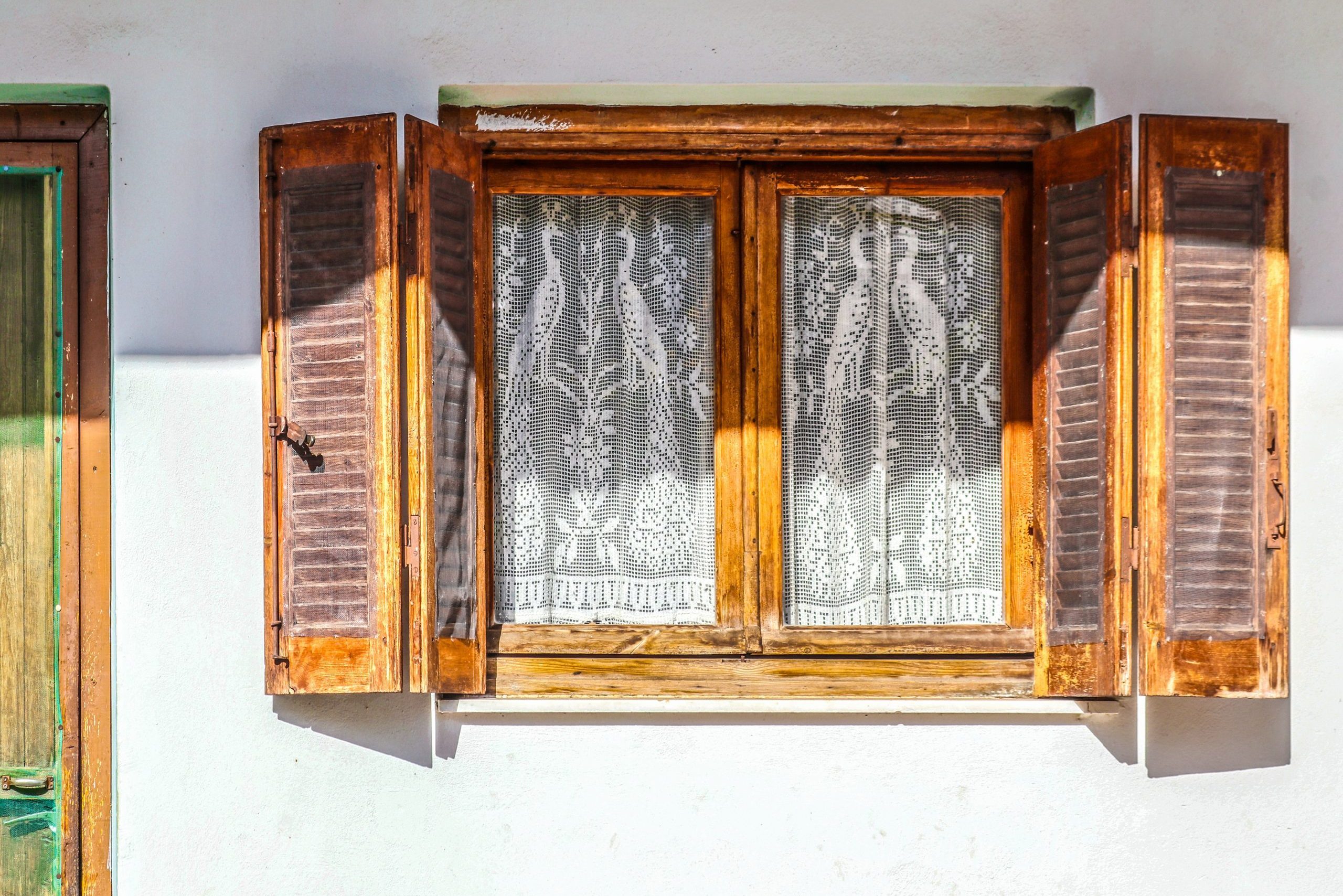 Old wooden window with open shutters on white stucco wall with insect netting tacked on and white lace peacock curtains - part of door visible - golden hour sun and shade