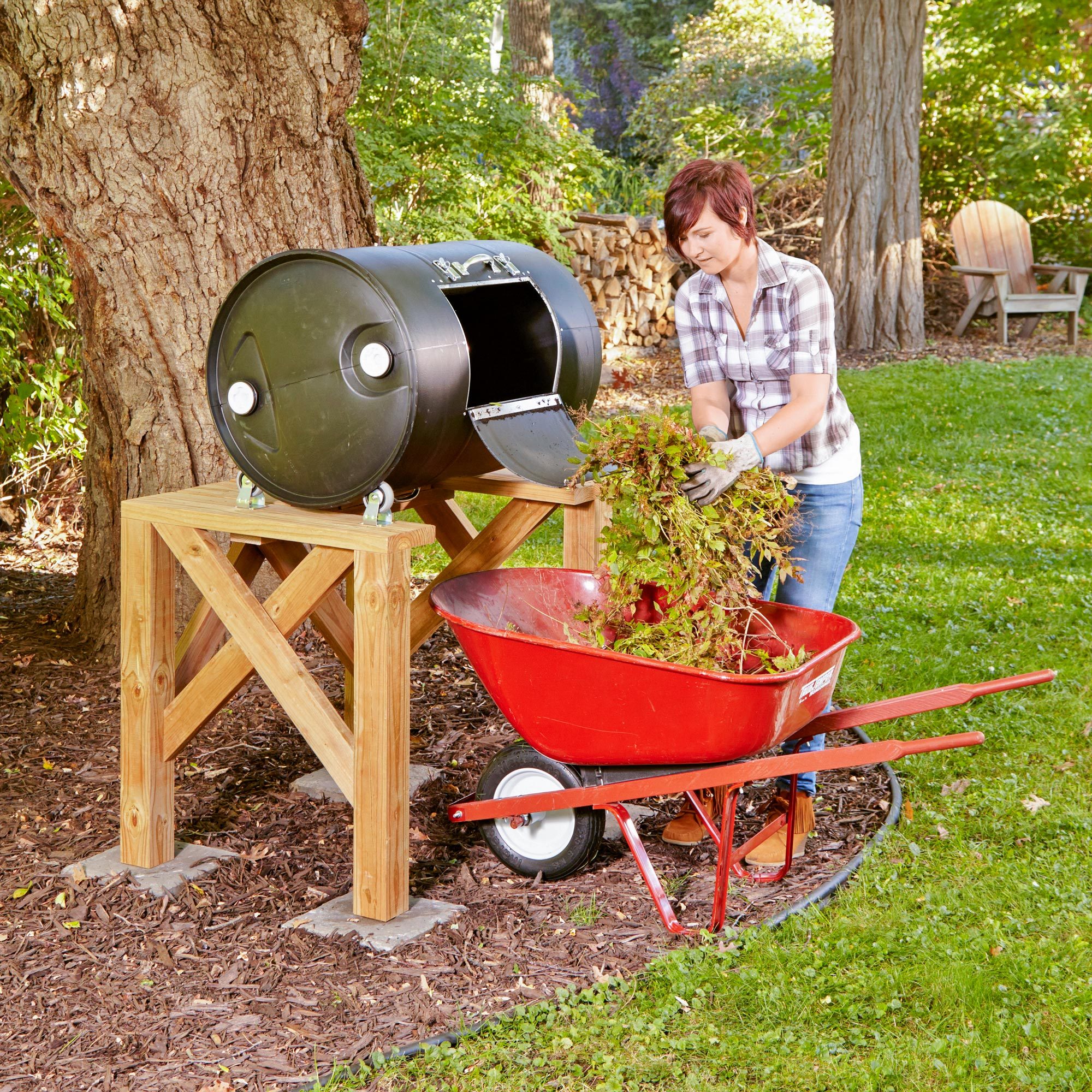 woman adding leaves from a whellbarrow to a homemade Drum Composter in a backyard under a large tree