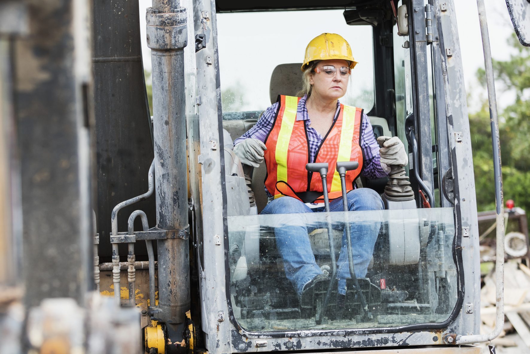 A mature Hispanic woman in her 40s working at a construction site, operating an earth mover. She is wearing a hardhat, reflective vest, and safety goggles.