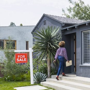 Female real estate agent carrying document while walking into house with a for sale sign out front