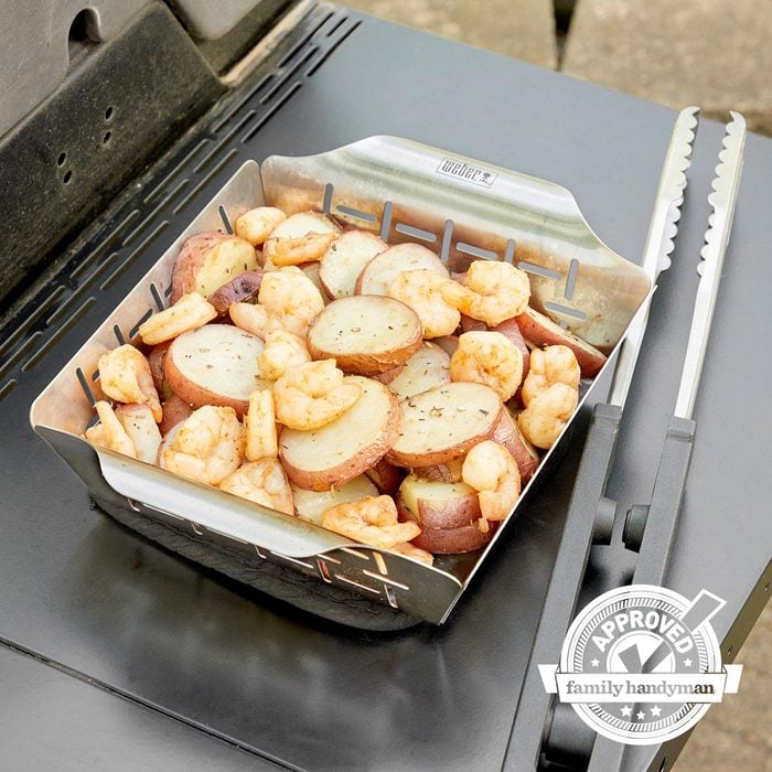 view from above of a weber grill basket with potatoes and shrimp next to a grill with tongs with the family handyman approved logo in the bottom right