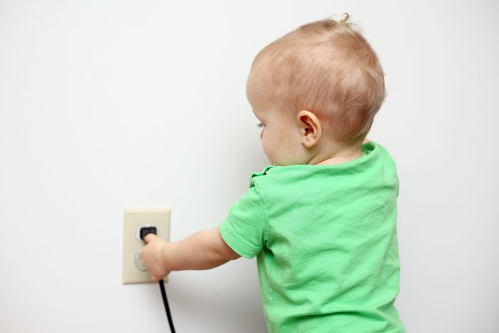 A close-up of a Caucasian baby boy standing near the electrical outlet and playing with a power cord plugged into it