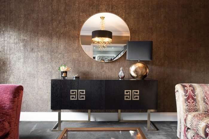 A Black Italianate and Greek style design wooden sideboard with brushed gold metal ornate Greek key-style handles and legs decorated with round gold table lamp with black shade, placed in front of a wall covered with brown textured wallpaper