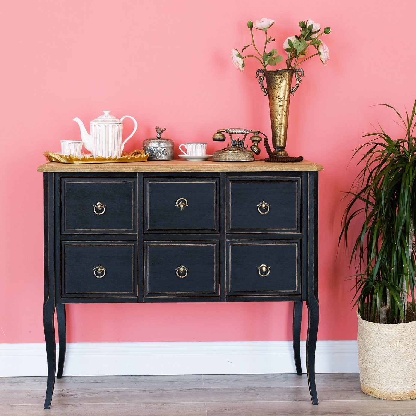 Old chest of drawers with objects on the background of a pink wall. Next is a flower in a pot