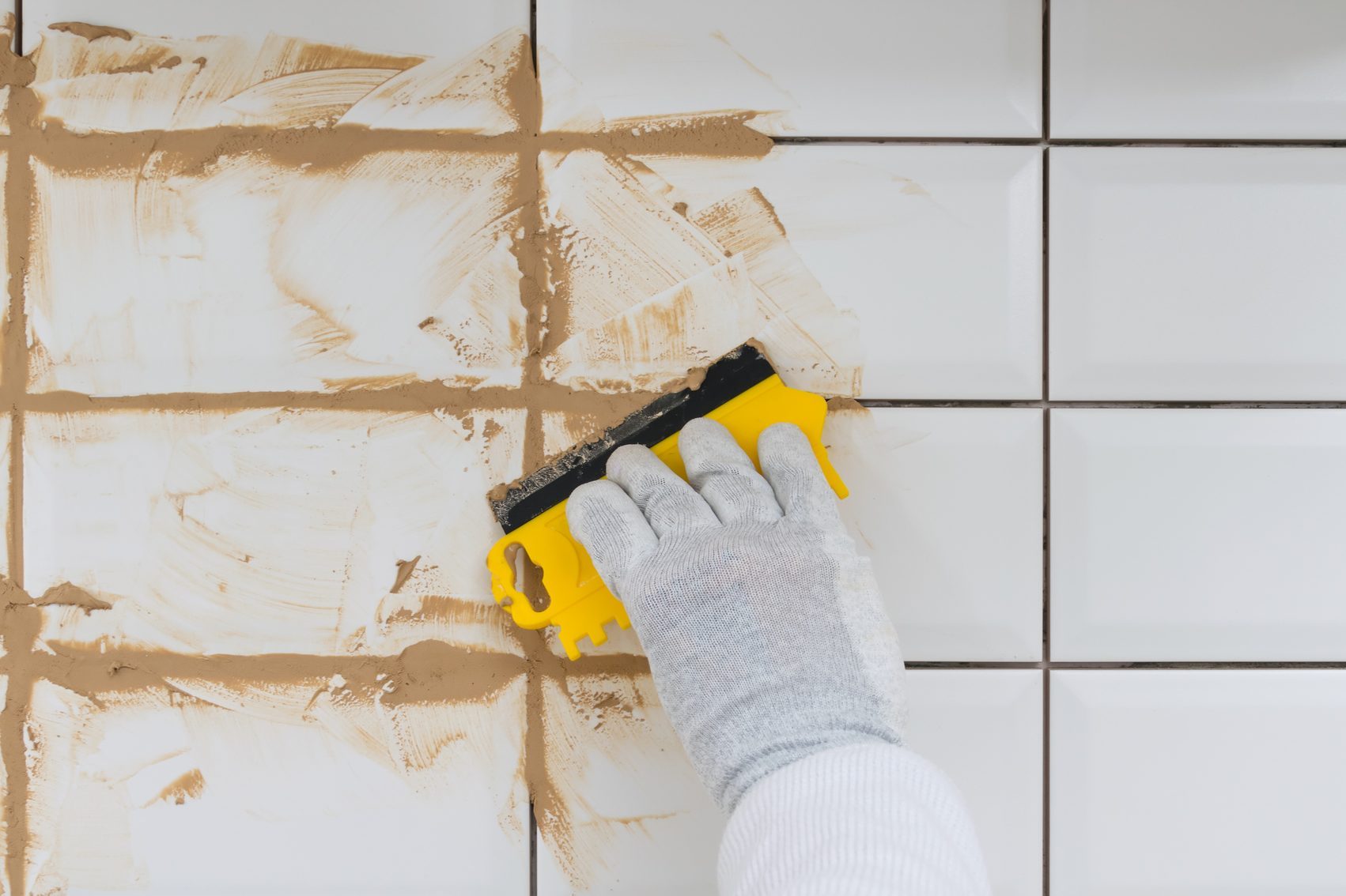 close-up of a hand in a protective glove holding a yellow spatula, in the process of brown grout on white tiles