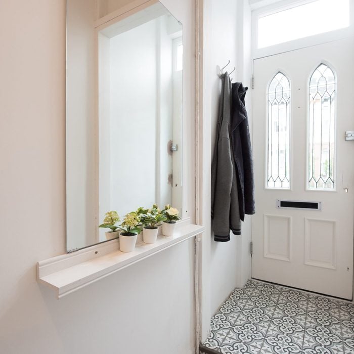 A general interior view of a hallway with a white front door, coats hanging from coat hooks, mirror above a shelf decorated with small artificial potted plants and a grey patterned tiled floor within a home