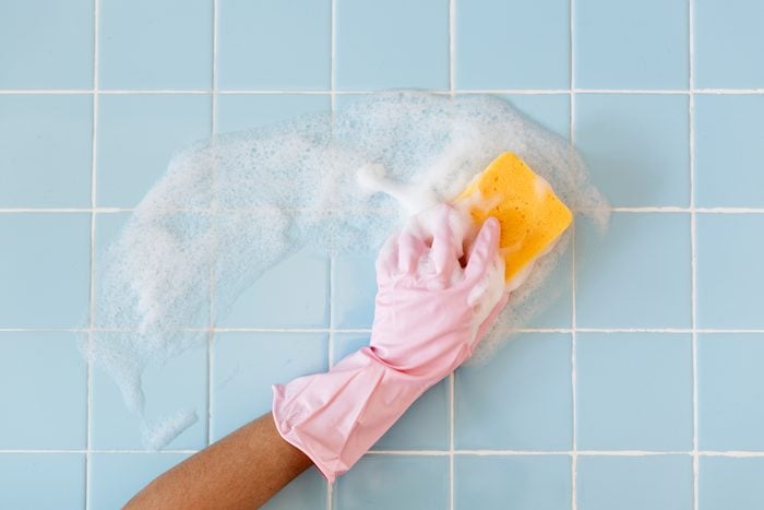 pink glove hand cleaning blue bathroom tile with a soapy yellow sponge