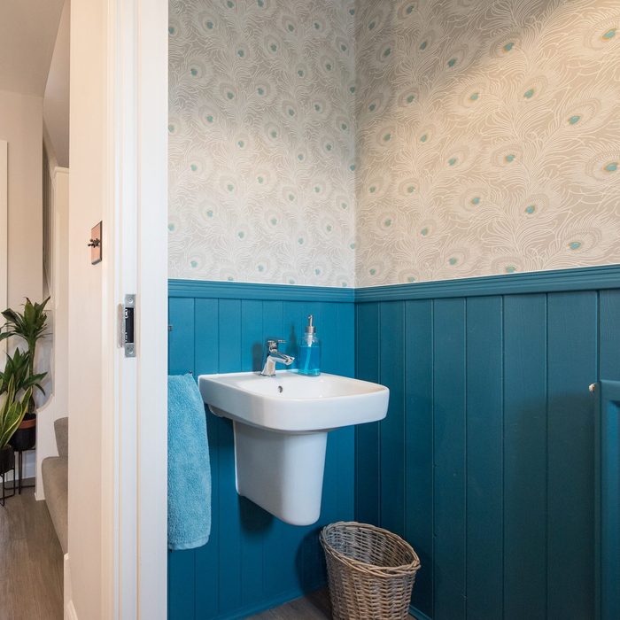 An interior view of a small downstairs toilet, with wood cladding, panelling painted blue and peacock feather pattern wallpaper looking through the hallway toward the lounge with vinyl wood effect flooring within a home