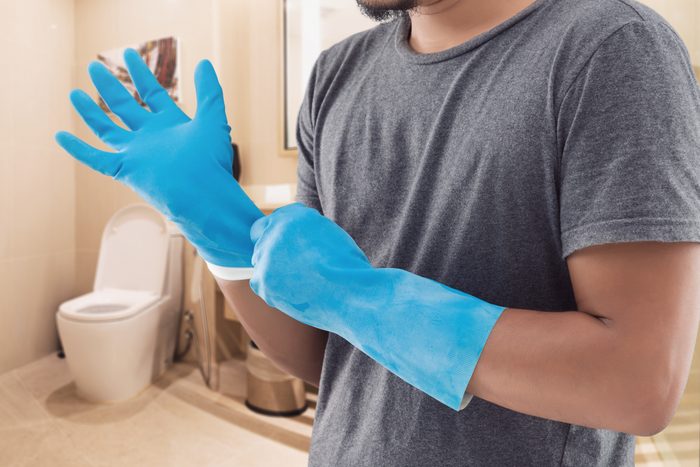 The Man In A Gray Shirt Wearing Blue Rubber Gloves Before Washing The Toilet.