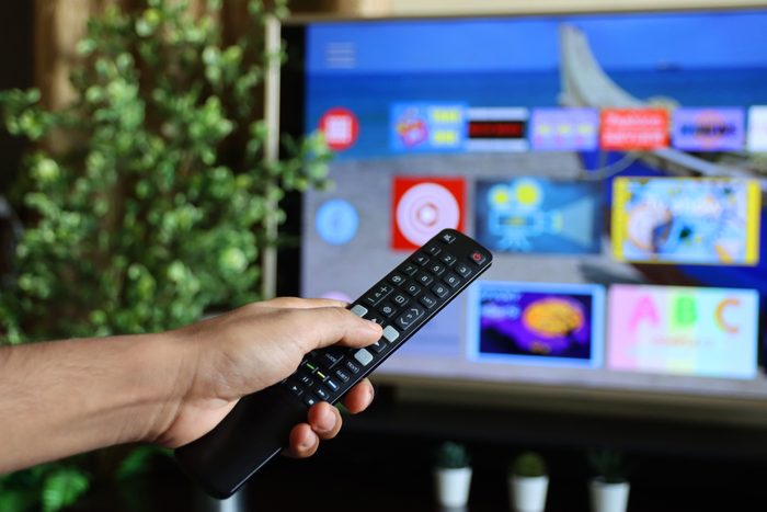 Image of unrecognisable person's hand holding a TV remote control, pointing at television screen, changing channels from rows of Smart Television Screen App Icons, background