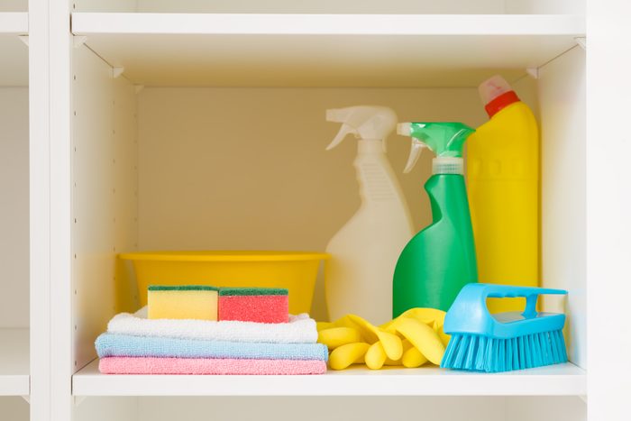 Bowl, brush, detergent bottles, rags, rubber gloves and sponges on white shelf inside opened wardrobe. Closeup. Set of colorful products for house cleaning. Front view.