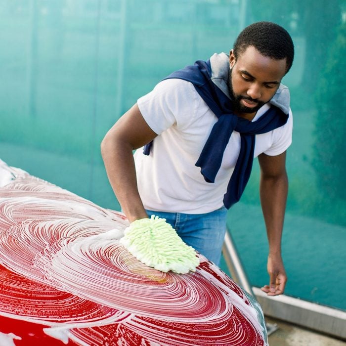 Horizontal view of car cleaning at self wash service outdoors. Handsome African man in casual wear washing red car with sponge and foam