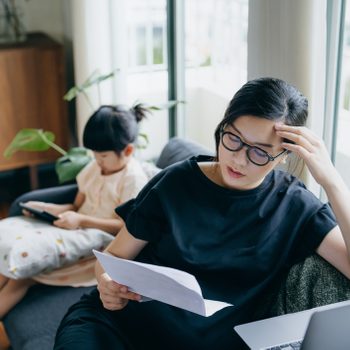 Young Asian mother looking stressed while managing her financial bills and tax documents, working from home on laptop and her daughter is using digital tablet in the background. Working mother managing work life and childcare at home