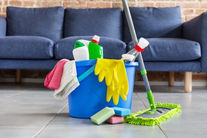 Bucket with bottles of detergent, cleaning tools and mopping stick in front of sofa in living room. Housework cleaning equipment.