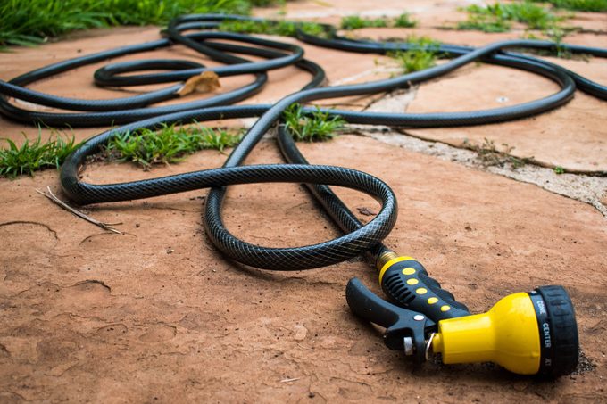 A Black Rubber Hose With A Bright Yellow Nozzle Randomly Coiled Across Terracotta Outdoor Tiles And Grass
