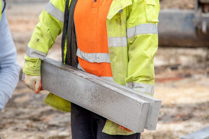 Close up of groundworker in orange and yellow hi-viz carrying heavy concrete kerbs on construction site during new road construction