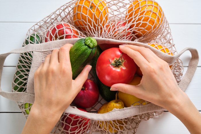 Fresh juicy fruits and vegetables in a reusable shopping bag. A girl or a woman takes or lays out products from a string bag made of recycled materials on a white wooden table or background. Vegetarianism, Veganism, Raw Food. Online shopping. No plastic.