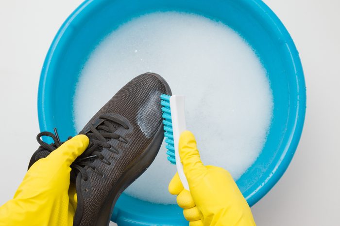 Hands in rubber protective gloves holding black sport shoe and brush above blue basin to wash the sneakers
