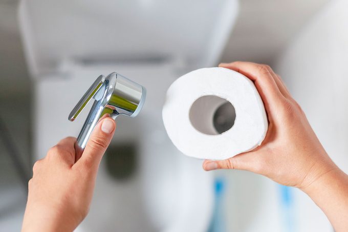 Man Holds Bidet Shower Wand and Toilet Paper above Toilet to determine which item to use