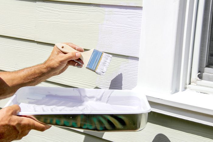 hand holding a paintbrush and paint tray painting the exterior of a house