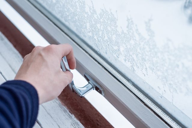 Person's hand crank opening window in the winter time with ice against the glass windows and snowy background