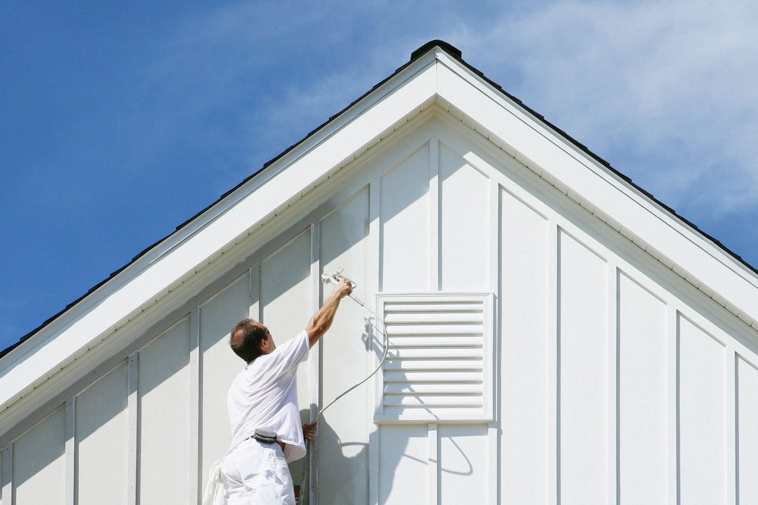 House Painter On Ladder Painting Home Exterior with a Paint Sprayer Gun and White Paint against a Blue Sky