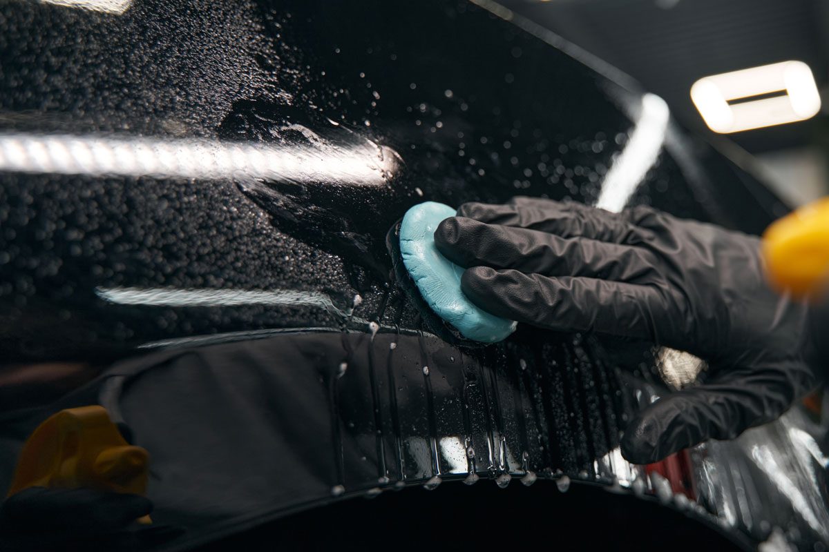 Cropped photo of skilled worker in nitrile gloves pressing lump of clay to car surface to remove paint impurities and to correct the paint to the correct color
